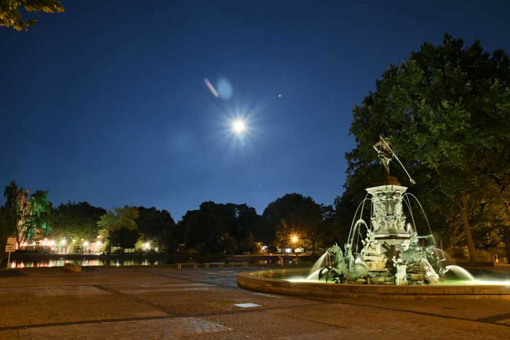 Nachtaufnahme des Nürnberger Stadtparks mit einem Brunnen, See und Restaurant im Hintergrund.